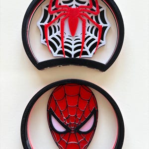 May include: Two circular ear-shaped accessories with black frames. The top piece features a red spider centered on a white and black web design. The bottom piece displays a red mask with black webbing lines and white eye lenses on a white background.