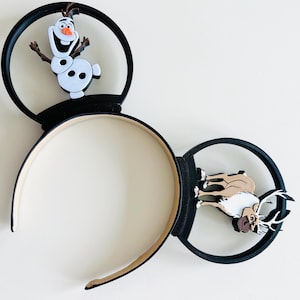 May include: Black mouse ears headband featuring Olaf the snowman and a reindeer. The Olaf figure is white with black eyes and a carrot nose. The reindeer is brown and white. The headband is beige and black.