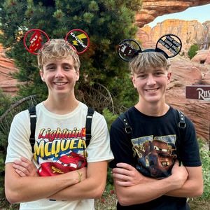 May include: Two young people wearing Disney-themed mouse ear headbands. One wears a white t-shirt with a Lightning McQueen graphic, the other a black shirt with a Mater graphic. Both are smiling with arms crossed.