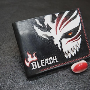 Puede incluir: Cartera de cuero negro con un gráfico blanco y rojo de la cara de un personaje de la serie de anime "Bleach". La cartera tiene costuras rojas y la palabra "BLEACH" en blanco con un diseño de llama roja. Una pequeña piedra roja y blanca está al lado de la cartera.