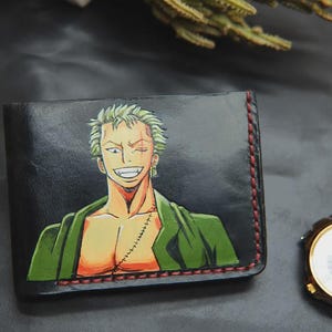 Könnte beinhalten: Schwarzes Lederportemonnaie mit einer farbenfrohen Illustration eines lächelnden Anime-Charakters mit grünen Haaren und einer Narbe. Das Portemonnaie hat rote Nähte an den Rändern. Eine goldene Armbanduhr mit braunem Lederarmband liegt neben dem Portemonnaie.