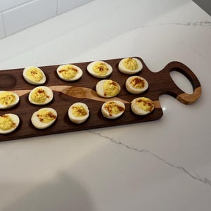 Puede incluir: Una tabla de servir de madera con asa, que contiene huevos rellenos. Los huevos están cortados por la mitad, rellenos con relleno amarillo y espolvoreados con pimentón. La tabla es de madera oscura con un acento de madera más clara.