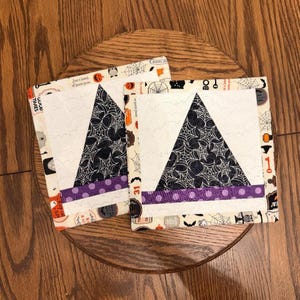 May include: Two square Halloween fabric coasters. Each coaster features a white triangle with a black spiderweb design, a purple polka dot stripe, and a patterned border with Halloween motifs. The coasters are on a wooden surface.