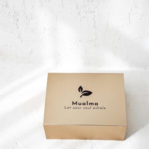 Könnte beinhalten: Eine goldfarbene Schachtel mit dem Text "Mualma" und "Let your soul exhale" in Schwarz. Die Schachtel hat ein einfaches Blattdesign über dem Text. Der Hintergrund ist eine strukturierte weiße Oberfläche.