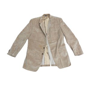 May include: A beige corduroy blazer featuring a notched lapel, two front buttons, and two chest pockets with pointed flaps. The jacket has long sleeves with buttoned cuffs and a light-colored interior lining visible through the open front.