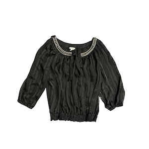 May include: Black blouse with vertical stripes and three-quarter sleeves. The neckline is embellished with small, silver-colored beads. The blouse has a tie at the neckline and a gathered waistline.