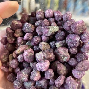 May include: A close-up of a pile of small, rough, purple ruby crystals. The crystals are in a clear glass jar.