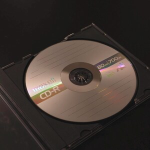 May include: A silver Maxell CD-R disc inside a clear plastic case. The disc has the text "maxell CD-R" and "80 700" printed on it. The case is open, and the background is dark.