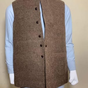 May include: A brown wool vest with a mandarin collar and a button-down front. The vest is worn over a light blue shirt. The vest has two pockets and is displayed on a mannequin.
