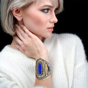 Può includere: Un bracciale a polsino dorato con dettagli intricati e pietre blu incastonate. Il bracciale presenta una pietra centrale ovale e due pietre più piccole sui lati. Il design ha un'estetica vintage o etnica.