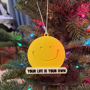 May include: A yellow smiley face Christmas ornament with the text "YOUR LIFE IS YOUR OWN" on a white banner. The ornament hangs on a green Christmas tree decorated with colorful lights.