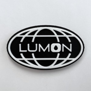 May include: Black and white oval-shaped emblem with the word "LUMON" in white, centered on a black background. The word is framed by a white globe design. The emblem is set against a plain white background.