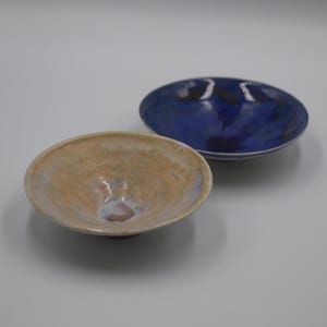 May include: Two shallow ceramic bowls with a smooth, glossy finish. One bowl features a light beige and tan glaze with subtle iridescent highlights, while the second bowl displays a deep, vibrant blue glaze with darker, mottled patches throughout.