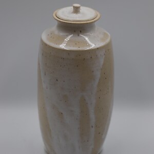 May include: A cylindrical ceramic jar with a lid, featuring a speckled tan base glaze overlaid with a white drip glaze that cascades down the sides. The vessel has a narrow neck, a flat rim, and a small knob handle centred on the circular lid.