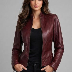 May include: A deep burgundy leather jacket featuring a mandarin collar, a full-length front zipper, and long sleeves. The jacket is worn over a simple black top and black pants, showcasing a fitted silhouette with visible seam detailing.