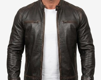 May include: A dark brown leather jacket with a distressed finish, featuring a stand-up collar with a snap button, a full-length front zipper, and two horizontal zippered chest pockets. The jacket is worn over a plain white t-shirt.