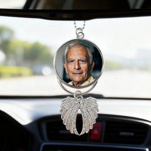 Op de afbeelding: Een auto achteruitkijkspiegel ornament met een cirkelvormige foto van een oudere man, opgehangen aan een ketting. Onder de foto een zilverkleurig engelenvleugelontwerp. Het ornament hangt voor het dashboard en de voorruit van een auto.