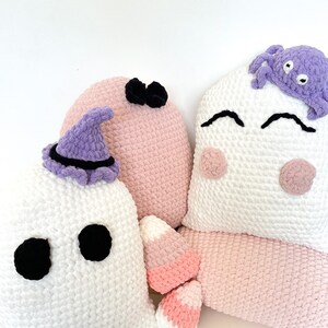 May include: Three crocheted Halloween cushions. One is a white ghost with black eyes, another is a pink cushion with a black bow, and the third is a white cushion with a purple crocheted spider on top.