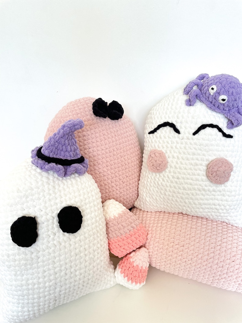 May include: Three crocheted Halloween cushions. One is a white ghost with black eyes, another is a pink cushion with a black bow, and the third is a white cushion with a purple crocheted spider on top.