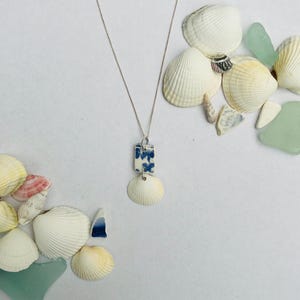 Puede incluir: Un collar de plata con un colgante rectangular floral azul y blanco y un colgante de concha blanca. El collar se muestra con conchas marinas y vidrio marino sobre una superficie blanca.