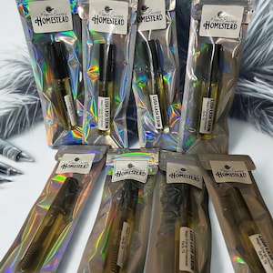 Op de afbeelding: Verschillende heldere glazen flesjes Lush Lash Serum zijn verpakt in iriserende, hersluitbare zakjes. Elk zakje heeft een wit etiket met de tekst "Magic Energy Homestead". De serumflesjes hebben zwarte doppen en zijn gevuld met een gouden vloeistof.