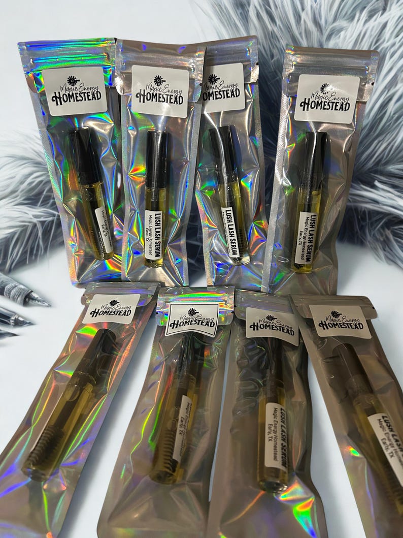 Op de afbeelding: Verschillende heldere glazen flesjes Lush Lash Serum zijn verpakt in iriserende, hersluitbare zakjes. Elk zakje heeft een wit etiket met de tekst "Magic Energy Homestead". De serumflesjes hebben zwarte doppen en zijn gevuld met een gouden vloeistof.