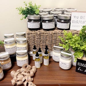Op de afbeelding: Een display van diverse huidverzorgingsproducten in glazen potten en flesjes, waaronder scrubs, boters en oli&euml;n. De potten zijn gelabeld en gerangschikt op een houten oppervlak met decoratief groen en borden met de tekst "You Are Amazing!" en "Human Kind be both."
