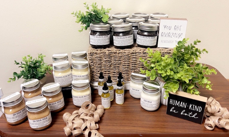 Op de afbeelding: Een display van diverse huidverzorgingsproducten in glazen potten en flesjes, waaronder scrubs, boters en oli&euml;n. De potten zijn gelabeld en gerangschikt op een houten oppervlak met decoratief groen en borden met de tekst "You Are Amazing!" en "Human Kind be both."