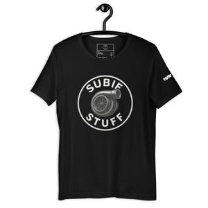 以下が含まれることがあります： ターボチャージャーと「SUBIE STUFF」の文字が描かれた白い円形グラフィックが特徴の黒い半袖Tシャツ。Tシャツは黒いハンガーにかかっています。