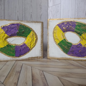 May include: Two rectangular white wooden plaques featuring a raised, textured king cake design. The cake is painted in alternating segments of yellow, purple, and green with gold accents. Each plaque has a gold-painted border and edges.