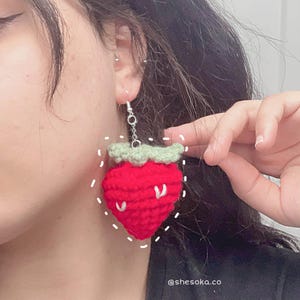 May include: A single red crochet strawberry earring with a green top and white seeds. The earring hangs from a silver chain and hook. The text "@shesoka.co" is visible at the bottom right.