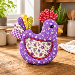 May include: A fabric chicken-shaped ornament featuring a purple body with a white daisy print, a red comb, and a multi-colored tail with polka dot, floral, and gingham patterns. The wing is a cream-colored patch with purple flowers and a red border.