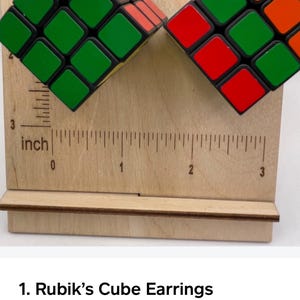 Puede incluir: Pendientes de cubo de Rubik con cuadrados verdes, rojos y naranjas. Los pendientes se muestran en un soporte de madera con una regla. Los pendientes miden aproximadamente 2,5 cm.