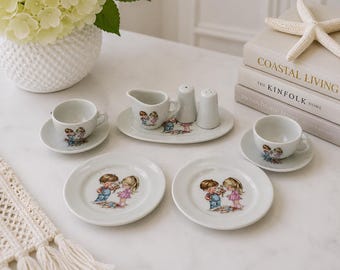 May include: A miniature porcelain tea set featuring two cups, two saucers, two plates, a creamer, and salt and pepper shakers. Each piece is white with a colourful illustration of a young boy and girl sharing flowers. The set is arranged on a surface.
