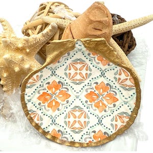 Op de afbeelding: Een decoratieve zeeschelp met een goudkleurige rand en een patrooninterieur met oranje bloemmotieven op een witte achtergrond. De afbeelding bevat ook zeesterren en andere natuurlijke elementen, die een kustthema oproepen.