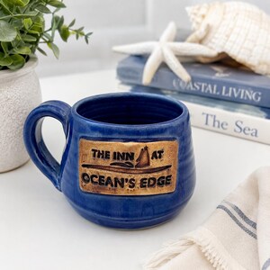 May include: A blue ceramic mug with a handle and a rectangular plaque. The plaque reads "THE INN AT OCEAN'S EDGE" with a sailboat illustration. The mug is sitting on a white surface. Coastal decor.