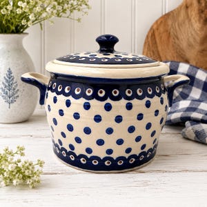 Puede incluir: Una olla de cerámica con tapa y dos asas laterales, con una base de color crema decorada con un patrón de lunares azul oscuro y bordes festoneados que contienen pequeños acentos circulares marrones y azules alrededor del borde y la base.