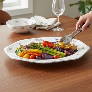 Könnte beinhalten: Eine weiße Servierplatte mit Blumendekor enthält geröstetes Gemüse, darunter Spargel, Karotten und Kartoffeln. Eine passende Schüssel und ein Weinglas stehen auf einem Holztisch. Eine Person serviert das Essen mit einer Zange.