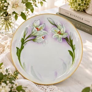 May include: A round white ceramic plate featuring a hand-painted design of two white lily stems with green leaves and red stamens against a soft purple wash background. The plate has a thin gold rim and a signature on the left side.