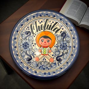May include: A decorative ceramic plate featuring a hand-painted design. The plate has a blue and white floral border and the word "Cholula" in black script. The center depicts a cartoon figure in traditional attire.