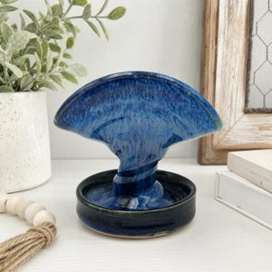 May include: A ceramic napkin holder featuring a deep blue glaze with lighter blue and white swirling patterns. The piece has a circular base and a curved, fan-shaped top section connected by a central twisted stem, creating a functional design.