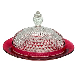May include: A clear glass butter dish with a ruby red base. The domed lid features a diamond pattern and a decorative finial. The base is a solid, circular plate. This elegant dish is perfect for serving butter.