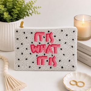 Könnte beinhalten: Eine rechteckige Reißverschlusstasche, die mit weißen Perlen und verstreuten schwarzen Perlen bedeckt ist. Der Text "IT IS WHAT IT IS" ist mittig in rosa Perlen gestickt. Die Tasche hat einen goldfarbenen Metallreißverschluss an der Ecke.