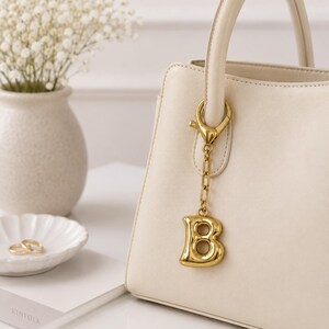 May include: A gold-toned metal letter B charm attached to a matching chain and lobster clasp, hanging from the handle of a cream-colored handbag. The charm features a smooth, rounded, and polished surface with a hollow center design.