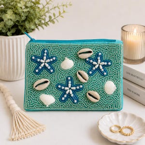 May include: A rectangular teal pouch featuring intricate beadwork in a sea-inspired pattern. The surface is adorned with three blue beaded starfish, white pearl accents, and several natural tan and white seashells attached to the fabric.
