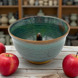 Può includere: Una ciotola per mele in ceramica fatta a mano con una glassa verde e turchese, con un supporto marrone per il torsolo della mela al centro. La ciotola ha una base gialla e un bordo marrone. Tre mele rosse sono sulla superficie di legno.
