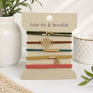 May include: A selection of hair ties and bracelets in shades of green, brown, teal, yellow, and coral. Each accessory features a gold-coloured accent. The card is labelled "hair tie & bracelet".