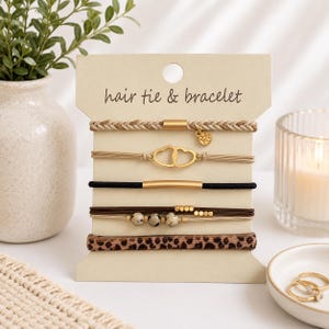May include: A set of hair ties and bracelets displayed on a beige card. The set includes a braided beige hair tie, a gold heart bracelet, a black hair tie, a brown bracelet with beads, and a leopard print hair tie. The card reads "hair tie & bracelet."