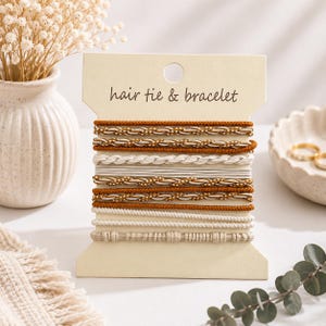 Puede incluir: Un conjunto de gomas para el pelo y pulseras presentadas en una tarjeta beige con la inscripción "hair tie & bracelet". La colección incluye varias texturas y colores, con bandas de color óxido, cordones dorados trenzados y lazos blancos trenzados.
