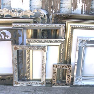 May include: A collection of various rectangular picture frames in metallic finishes including silver, gold, and bronze. The frames feature diverse decorative borders with intricate scrollwork, floral patterns, and smooth edges, arranged in a layered group.